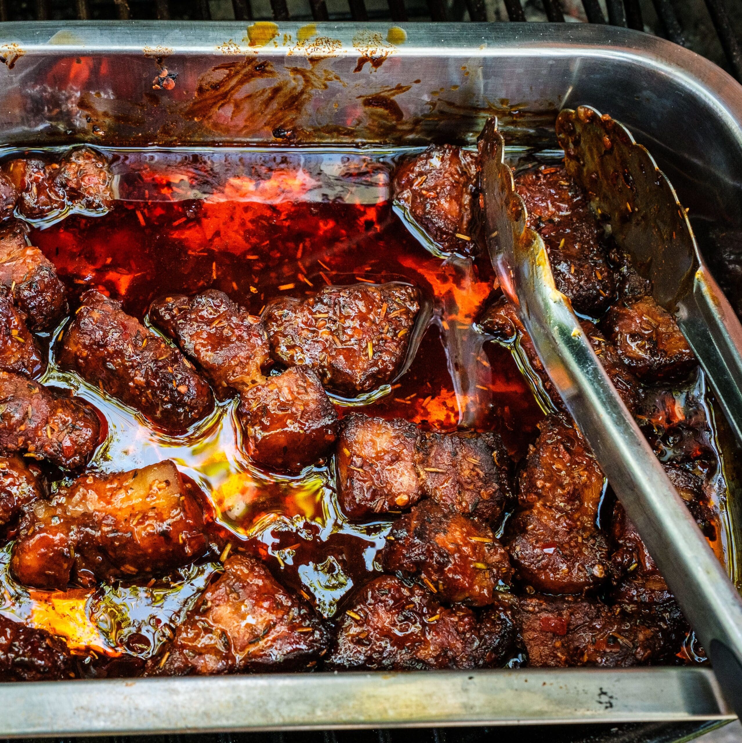 A metal pan on a grill with burnt ends cooking.